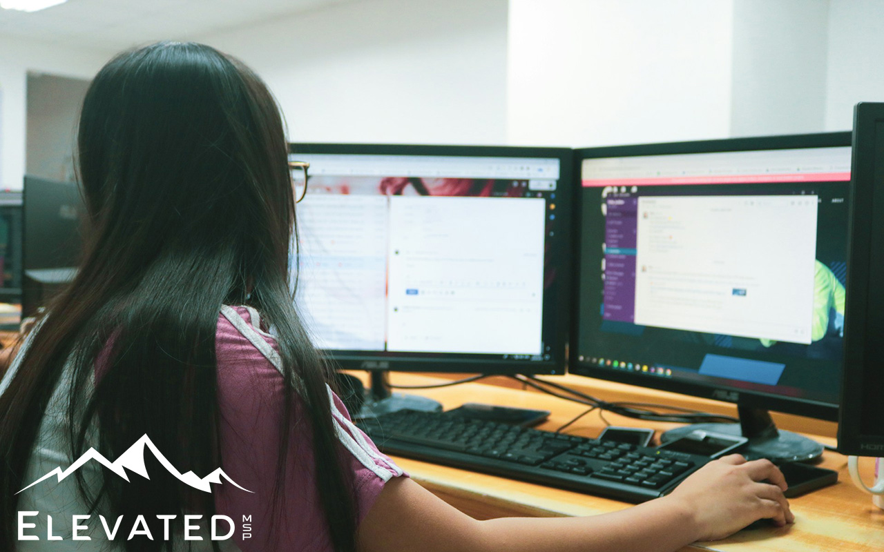 Woman with long black hair sitting in front of dual computer monitors with her hand on her mouse.