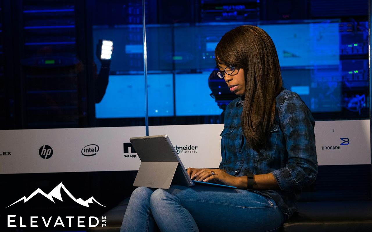 Long haired black female, wearing a green and black checked flannel shirt, sitting in front of a server room with a laptop open on her lap.